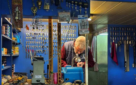 Locksmith working at a bench in a key shop surrounded by hanging keys and tools