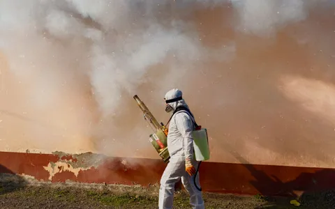 Pest control technician in a white protective suit and respirator fogging the exterior of a property